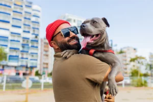 Dog walker wearing sunglasses and a beanie holds a pitbull wearing a harness outdoors in a city setting.