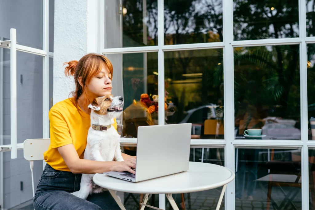 Pet professional working on laptop at outdoor cafe playfully cuddles a small dog